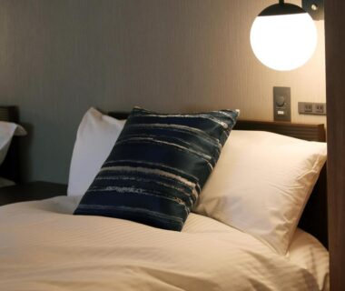 White bedding with navy striped cushion and round bedside lamp in a Hotel Ichibokaku guest room, Nagano