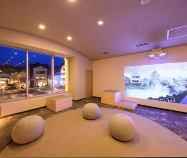 Interior lounge with curved window, low round seating and large projected screen overlooking Gunma town