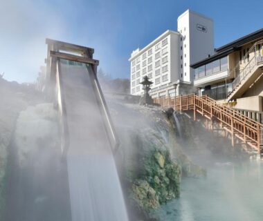 Steaming hot spring cascade flowing into onsen pool with wooden walkways and hotel building in Gunma
