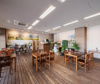 Communal dining area with wooden floors, tables, chairs, plants and shelving at Hotel Kasuien in Saga