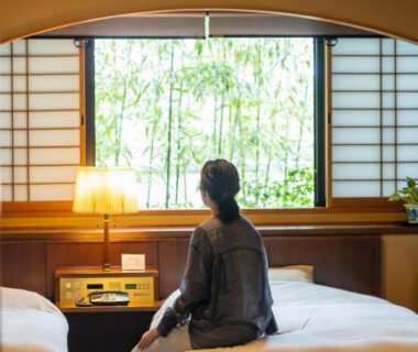 Woman seated on twin beds beside shoji window with bamboo outside at Hotel Kazahaya, Ōita