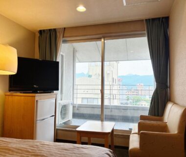 Compact guest room with bed, TV, small fridge, seating area and balcony overlooking city and distant mountains