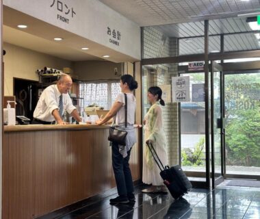 Front desk staff assisting guests with luggage beside glass entrance with greenery outside