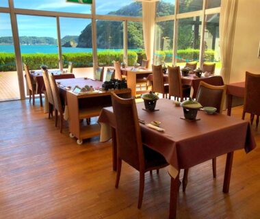 Bright dining room with wooden floors and tables set by large windows overlooking water and hills