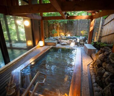 Indoor wooden onsen pool with stone edging, lantern lighting and large windows overlooking lush greenery at Hotel Nanpuso Kanagawa