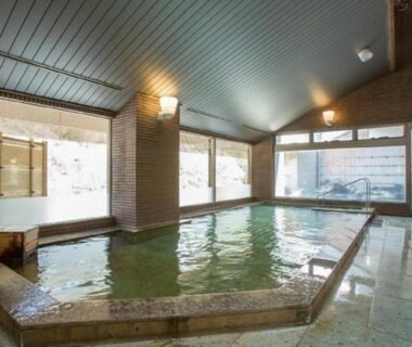 Spacious indoor onsen with stone-bordered bath, tiled floor and large windows at Hotel New Momiji in Tochigi