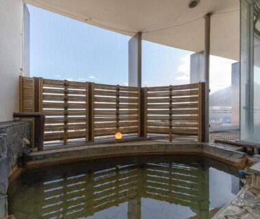 Round wooden soaking bath with stone edge and slatted wooden fence at Hotel New Tsuruta, Ōita