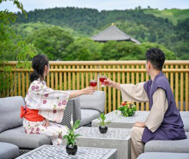 Couple in yukata clinking glasses on Hotel New Wakasa terrace with garden and distant temple in Nara