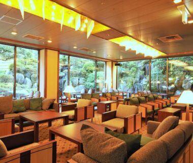 Spacious ryokan lounge with low wooden tables, upholstered sofas and large windows overlooking a manicured garden in Kanagawa