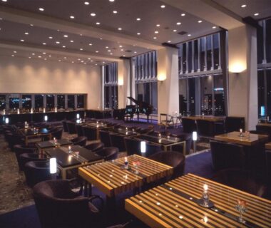 Spacious hotel lounge with wooden slatted tables, armchairs, grand piano and floor-to-ceiling windows overlooking city lights