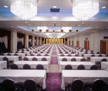 Carpeted banquet hall with rows of white tables, chairs, chandeliers and front stage at Hotel Ravie Kawaryo, Shizuoka