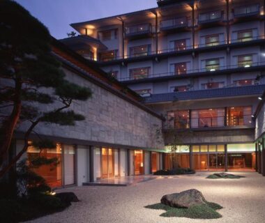 Evening view of a gravel courtyard garden with stone and pine in front of Hotel Saginoyu's lit facade, Nagano
