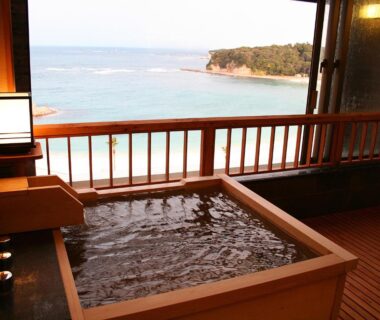 Wooden soaking tub on balcony with panoramic sea view and coastal cliffs beyond
