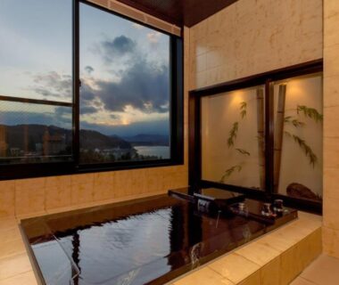 Tiled indoor onsen with wooden tub and large window framing distant mountains at dusk