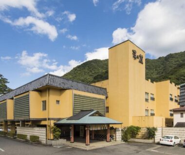 Yellow ryokan facade with covered entrance and mountain backdrop, Hotel Shirakawa Yunokura, Tochigi