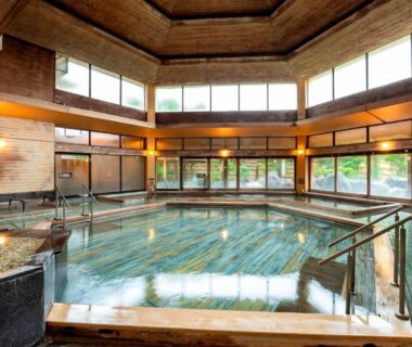 Spacious indoor onsen bath with wooden decking, handrails, large windows and garden views at Hotel Sunvalley Izunagaoka, Shizuoka