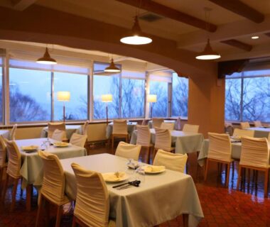 Bright ryokan dining with tables, white linens, chairs and large windows overlooking wooded mountain slopes in Nagano