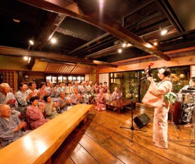 Guests in yukata watching kimono performer and band in a wooden ryokan communal hall
