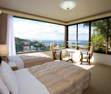 Twin bedroom with large corner windows overlooking coastline, chairs and low table, soft natural light