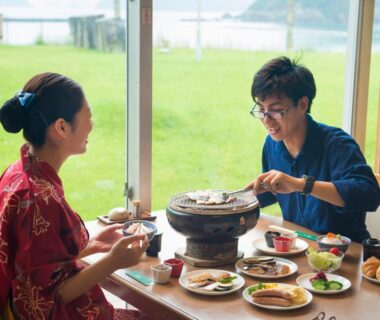 Two guests in yukata sharing a tabletop grill meal by window with grassy shore and water beyond