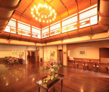 Spacious high-ceiling lobby with chandelier, polished wooden floors, reception counters and central flower arrangement at Hotel Tsubakino Nagano