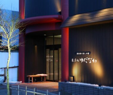 Evening entrance with illuminated signage, wooden slat facade and small tree at Hotel Yudanaka in Nagano