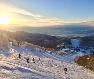 Skiers descending a snowy slope at sunset with resort buildings and mountain valley near Yudanaka, Nagano