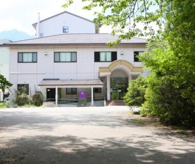 White three-story ryokan with arched entrance, gravel courtyard and surrounding trees near Nagano mountains