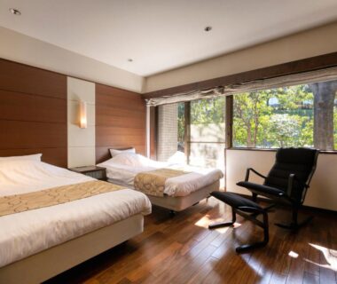 Sunlit twin beds with wooden floor and seating by a large window overlooking greenery at Hotel Yumeshizuku Kumamoto