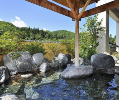 Open-air onsen bath with stone edging and wooden shelter overlooking green hills at Hotel Yumoto, Niigata