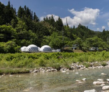Two white glamping domes beside a clear river with forested hills near Hotel&Glamp YAMABOSHI in Nagano