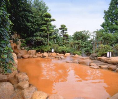 Rock-lined outdoor onsen with reddish water surrounded by trees and lanterns at Hyoe Koyokaku, Hyōgo