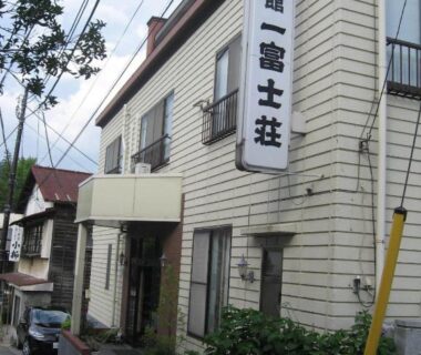 Two-story ryokan exterior with vertical signboard, entrance awning and potted plants along a narrow street in Yamanashi