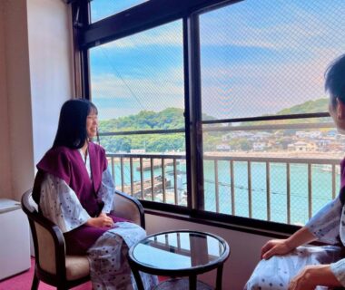 Two guests in robes seated by window overlooking town and waterfront from Ichifukuso in Saga