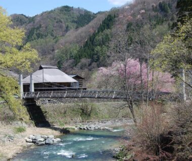 Wooden footbridge over river leading to traditional ryokan building amid mountains and spring blossoms in Nagano
