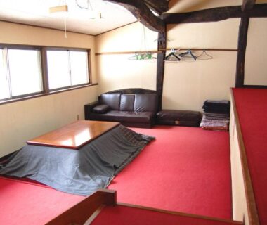 Low kotatsu table on red carpeted floor, sofa, folded futon stack and exposed wooden beams in Ichinokurasou Nagano room