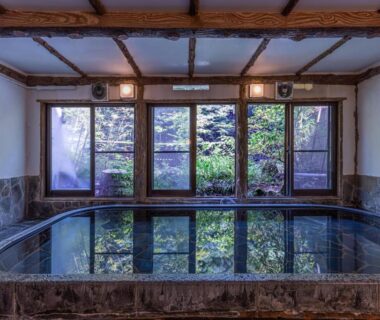 Stone-lined indoor onsen reflecting garden through wooden-framed windows at Ichinoyu Honkan, Kanagawa