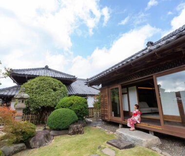 Guest in kimono on engawa overlooking manicured Japanese garden and traditional wooden ryokan buildings in Tochigi