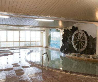 Spacious indoor onsen bath with tiled floor, waterwheel feature and large windows at Ikaho Onsen Todoroki, Gunma