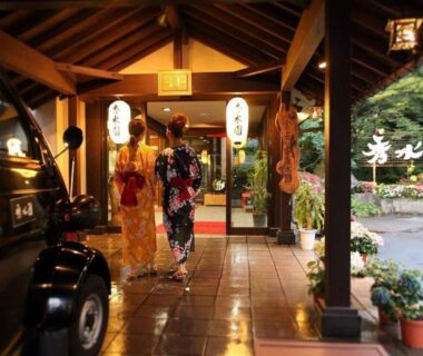 Two guests in yukata walking under a wooden covered entrance with lanterns and potted plants at Ikaho Syusuien, Gunma
