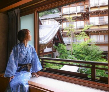 Guest in blue yukata seated by window overlooking traditional wooden ryokan exterior and small garden in Nagano