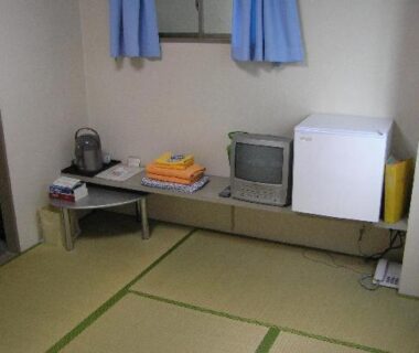 Compact tatami room with low table, small TV, mini fridge and stacked linens at Ikawa Ryokan in Hiroshima