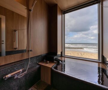 Wood-paneled bath with soaking tub and shower, window overlooking sandy beach, Ikkyu Seaside Hotel Yamagata