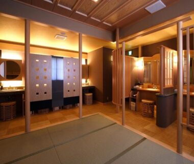 Warm-lit communal bathing changing area with tatami mat, wooden partitions, sinks and stools at a Shizuoka ryokan