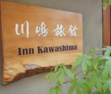 Wooden signboard with Japanese characters and Inn Kawashima text beside green foliage, exterior ryokan signage in Kyoto