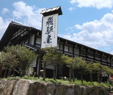 Traditional timber ryokan exterior with tall kanji sign, stone wall and landscaped shrubs at Iroriyado Hidaya, Gifu