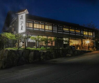 Evening lit two-story wooden ryokan facade with stone retaining wall and exterior lighting at Iroriyado Hidaya, Gifu, Chūbu