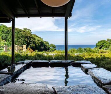 Outdoor stone onsen framed by wooden roof with sea and coastal greenery beyond at Isaribi, Shizuoka