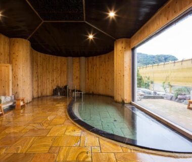 Indoor onsen with wooden walls, tiled floor, washing stations and large window overlooking outdoor terrace and hillside