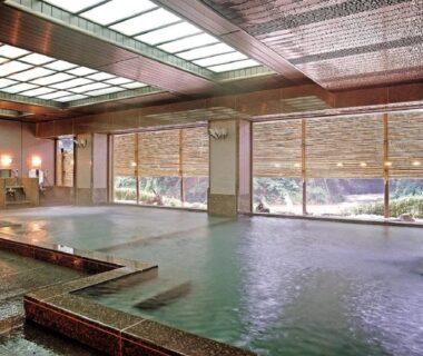 Spacious indoor communal onsen with stone-bordered pools, wooden blinds and skylight at Itoen Hotel Iizakakanouya Fukushima